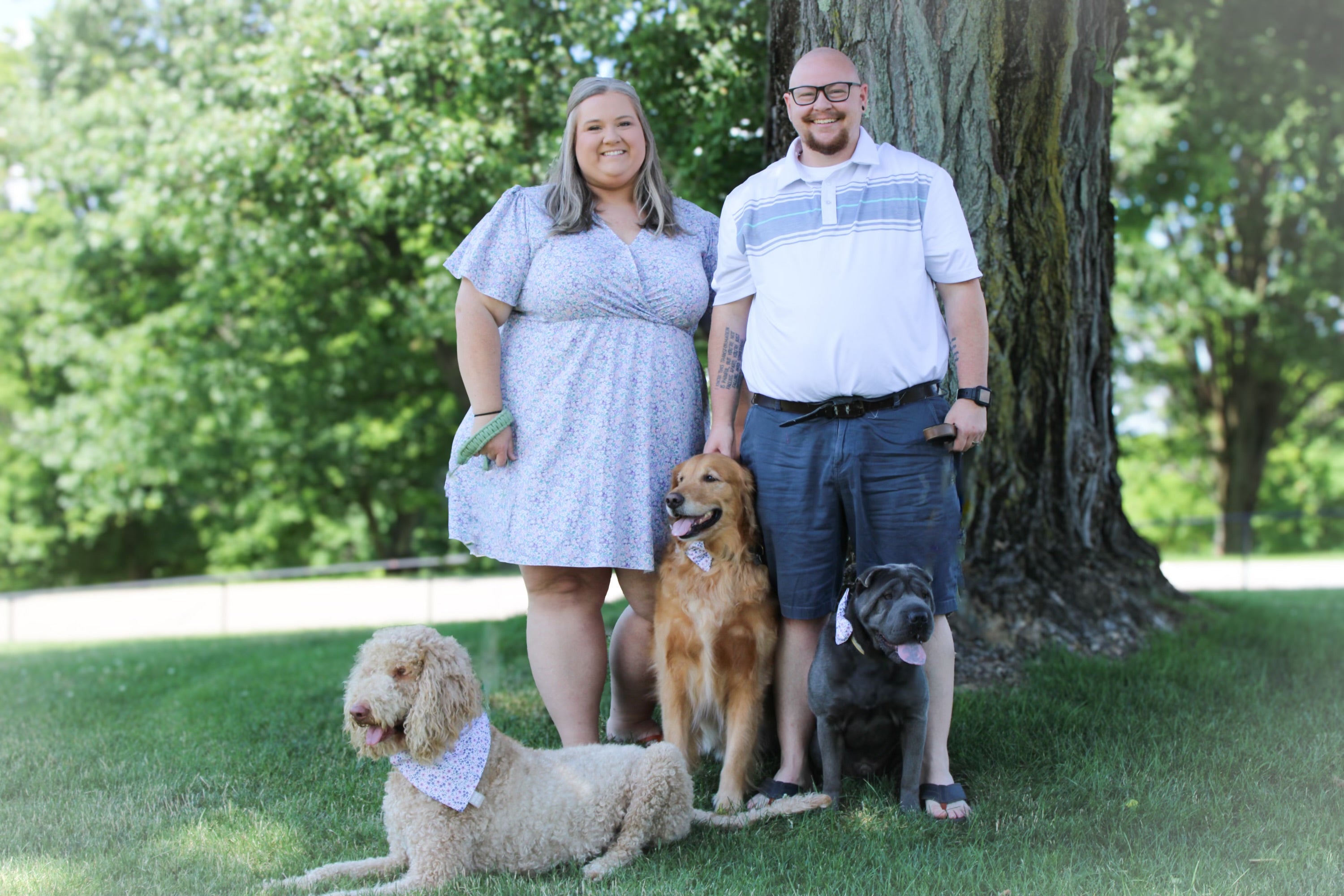 Two people and three dogs standing outdoors in a park-like setting with trees and grass.